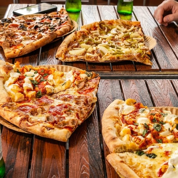 Four large pizzas with various toppings and beer bottles on a wooden table with people's hands.
